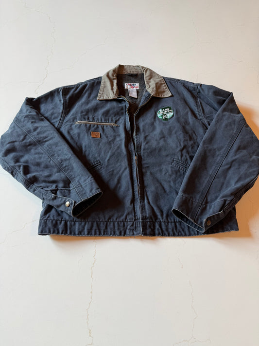 A vintage dark gray work jacket with a brown collar, laid flat on a light beige surface. The jacket features a zippered front, a chest pocket with a leather patch, and a circular embroidered patch on the left chest.
